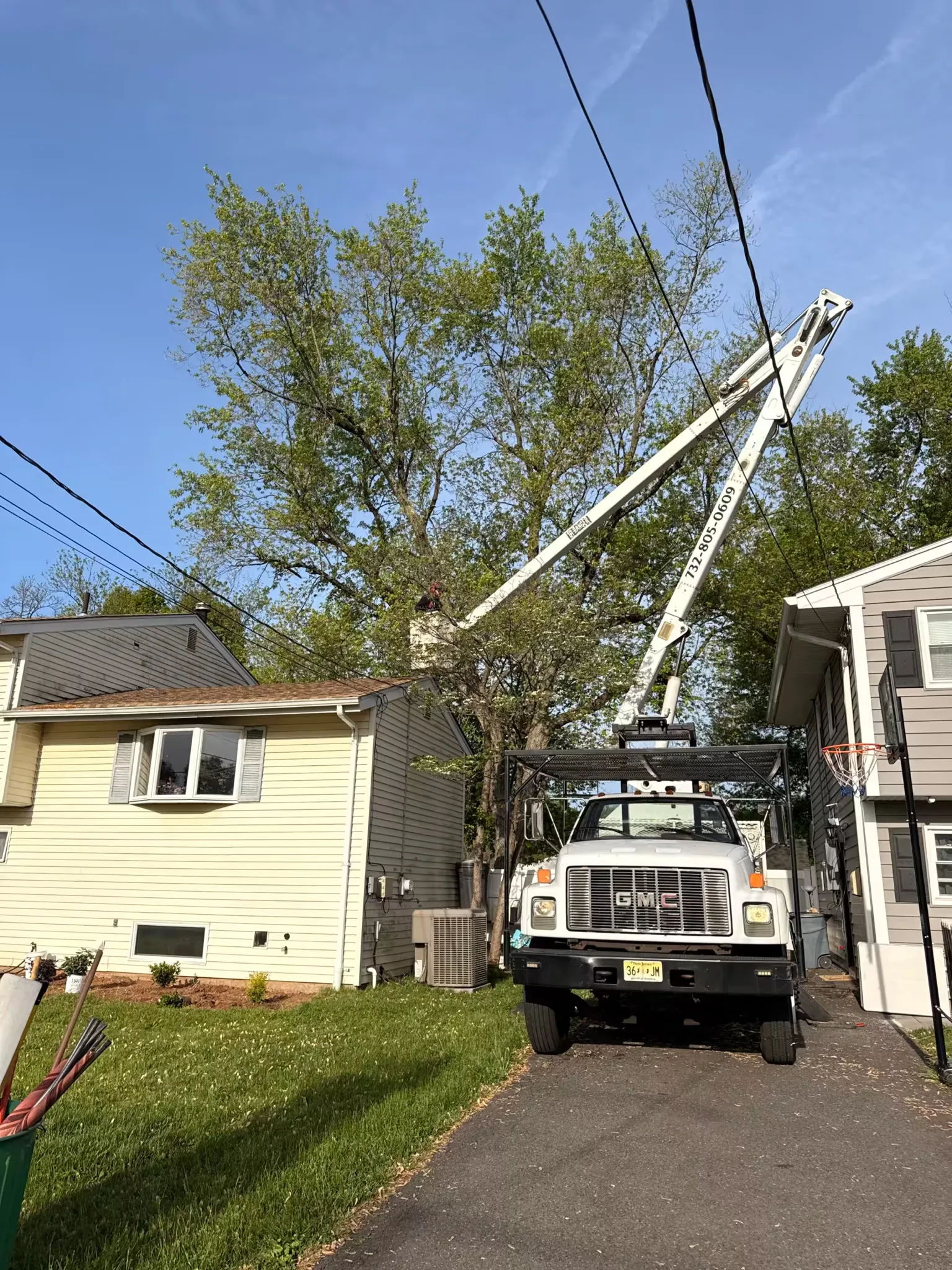 A white GMC boom truck parked in a driveway with an operator in the aerial lift bucket pruning tree branches over a house roof.