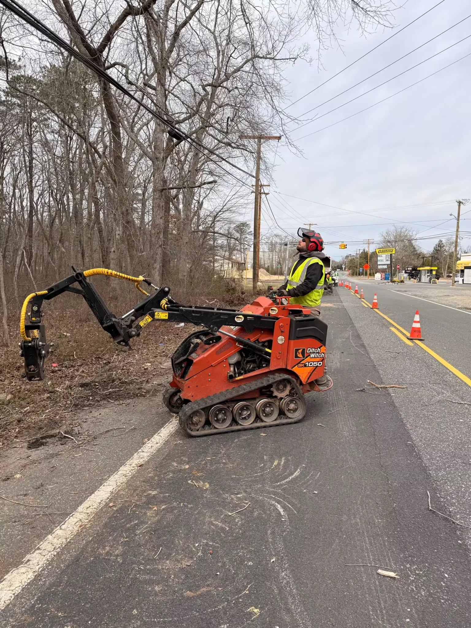 A worker wearing safety gear operates an orange Ditch Witch mini track loader with a grapple attachment along a roadside tree line.